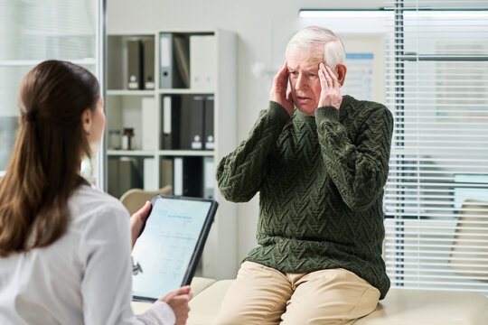 Senior man sitting on examination table holding head with both hands appearing to describe symptoms while young woman doctor holding clipboard listening attentively