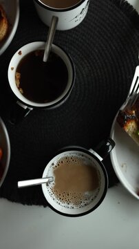 A table shows several mugs with different types of drinks. Plates hold slices of bread, vegetables, and sausages. It is morning time as people enjoy breakfast together.