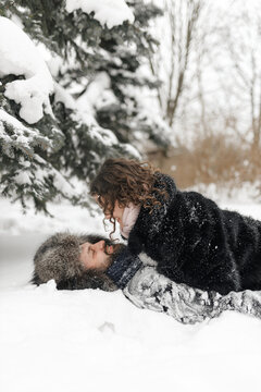 Side view of happy couple in winter clothes lying on snow in forest while having fun and looking at each other 