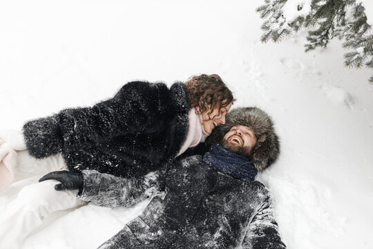 Playful couple in warm clothes lying on snowy ground in winter park and enjoying weather 