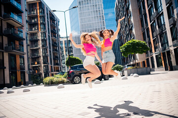 Two young girlfriends jump in the city street wearing bright outfits enjoying sunny day urban fashion and friendship while exploring modern city life and feeling joyful energetic and carefree together