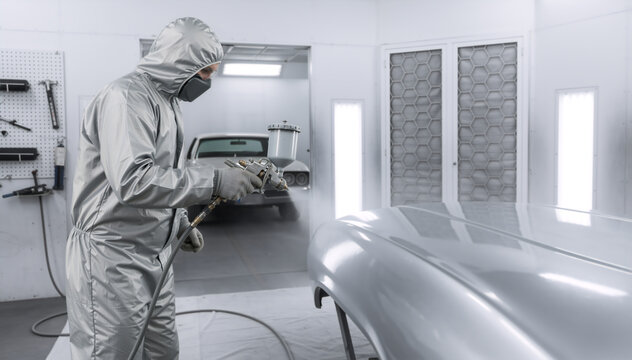 Auto body technician in protective suit spray painting a car panel. Professional mechanic using a paint gun in a brightly lit automotive repair booth