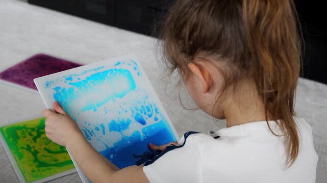 Little girl holding colorful sensory gel mat and pressing surface with finger while drawing shapes. Tactile stimulation and sensory development concept for autism support and occupational therapy.