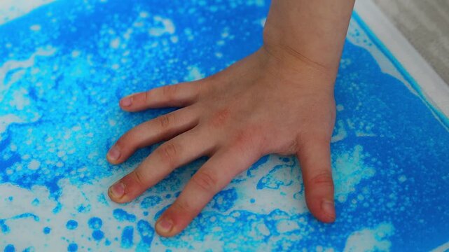 Child hand pressing colorful sensory gel mat creating moving patterns during tactile play. Sensory stimulation and occupational therapy concept for autism support and sensory integration development
