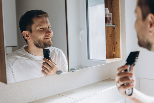 Man using electric razor for beard shaving in home bathroom mirror