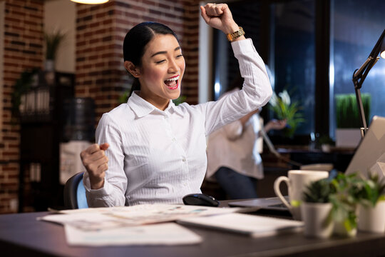 Happy asian entrepreneur celebrates a win at work, raising her arms in excitement while looking at her laptop. Cheerful businesswoman smiles brightly, feeling accomplished during her night shift.