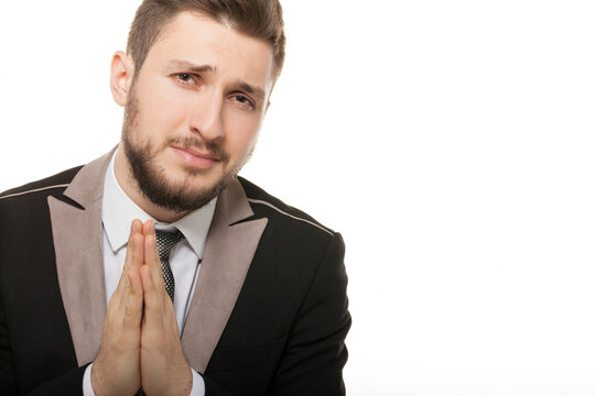 Sad young businessman with hands joined in prayer gesture, portrait of apologetic man in suit pleading for help or asking for forgiveness, isolated on white background with copy space.