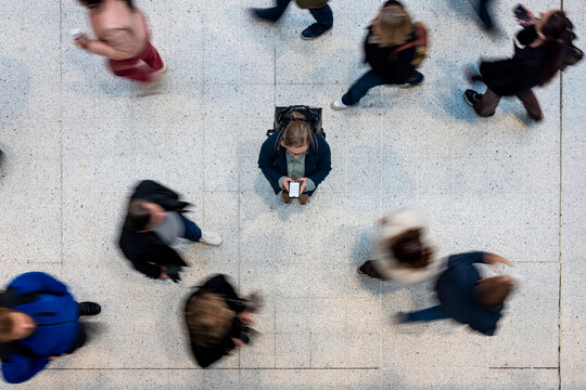 Commuter woman standing still with smartphone amidst rushing crowd overhead