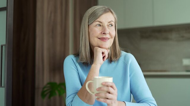 Happy smiling senior gray haired female relaxes holding cup of hot drink while sitting in kitchen at home. Calm satisfied elderly retired woman enjoying coffee or tea, feeling cozy and calm.