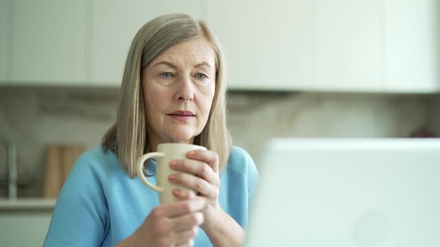 Senior gray haired female with mug uses computer sitting at kitchen at home. Elderly retired woman watching video news, browsing social media surfing web internet, online shopping, chatting. Close up.