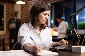 Closeup of caucasian woman working at her desk, focused on taking important notes on a notebook while using her laptop. The dim lighting highlights her night shift office environment. © DC Studio