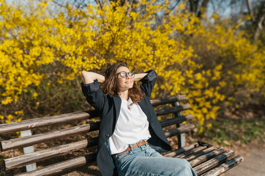 Relaxed young curly woman in glasses enjoying spring sun on a city bench under blooming yellow forsythia bushes