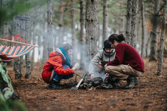 Three friends sit around a small campfire in a pine forest on a mountain. The group tends the fire and prepares for a cozy outdoor evening together.