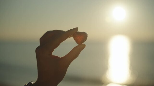 A young woman by the sea at dawn holds a heart-shaped chocolate in a red wrapper in her hand, close-up against the background of the sun's rays on the water.