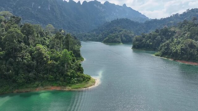 Aerial view of Khao Sok lake surrounded by lush green rainforest and dramatic limestone mountains under a cloudy sky, Khao Sok, Surat Thani, Thailand.