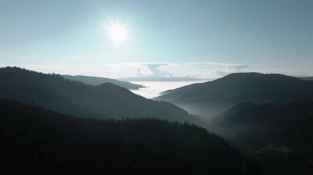 Aerial pull back over dark silhouetted mountains of Bicaz, Romania, revealing a thick sea of fog in the valley under a bright morning sun