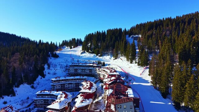 Ski resort is visible with buildings lined up at the foot of the slopes. Ski trails are marked on the snow-covered hills. Pine trees surround the area under bright sunlight.