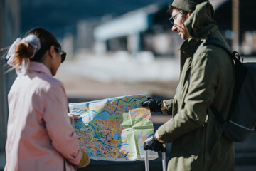 Obraz premium Two tourists examine a folded city map while standing with luggage outdoors. A couple studies directions together as they plan their travel route.