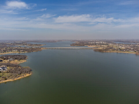 Aerial view of a vast lake reflecting the soft sky, divided by a long, straight bridge connecting tree-lined shores, Little Elm, Texas, United States.