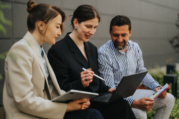 Three professionals collaborate while reviewing a laptop and tablet during an outdoor meeting. A...