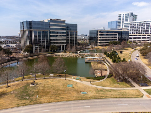 Aerial view of glassy buildings reflecting the serene lake, with pathways winding through the park, creating a tranquil urban oasis, Frisco, Texas, United States.
