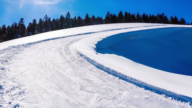 Curved Groomed Ski Slope Under Blue Sky, Crisp Sunlight Casting Long Shadows Across Compact White Snow, Pine Tree Line On Horizon, Empty Resort Run Offering Pristine Carving Surface And Wide