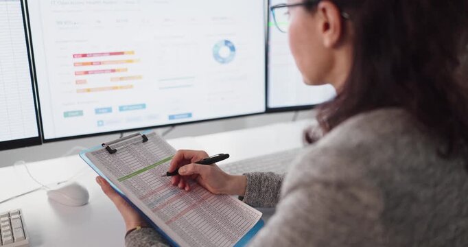 Woman accountant checks spreadsheet data report on computer. Close view shows analyst marking rows on clipboard while monitors show office finance. Clear audit data concept for finance.