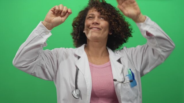 Woman doctor in white coat has fists raised and eyes closed in studio against bright green background celebrating a victory; joy.
