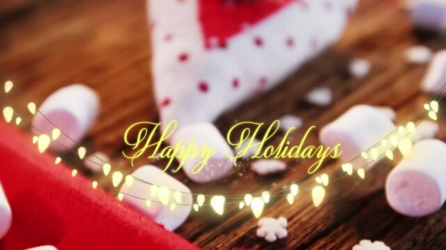 Animation of text, happy holidays, in yellow, over string lights, marshmallows and xmas decorations