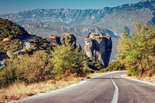 View of a winding road leading toward the majestic Meteora monasteries perched atop towering rock formations against a backdrop of mountains, Kalabaka, Greece.