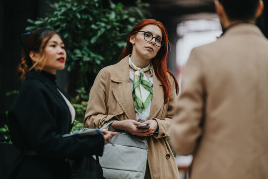 A group of business workers stand outdoors, with a red-haired woman holding a phone. Colleagues converse in an urban setting during a brief casual meeting.