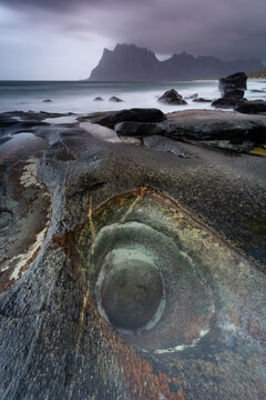 View of a textured rock formation with a circular hole meets the moody sea, framed by distant mountains under a stormy sky, Lofoten Islands, Nordland, Norway.
