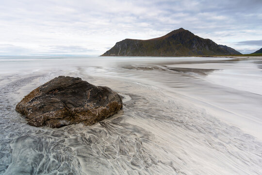 View of a solitary rock sits steadfast on the beach, the sand swirling around it like a silver river meeting the mountain backdrop, Lofoten Islands, Norway.