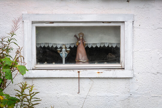 View of a white-framed window displaying a doll, a candle holder, and a lace curtain, set against a stark white wall contrasting with the greenery outside, Lofoten Islands, Norway.