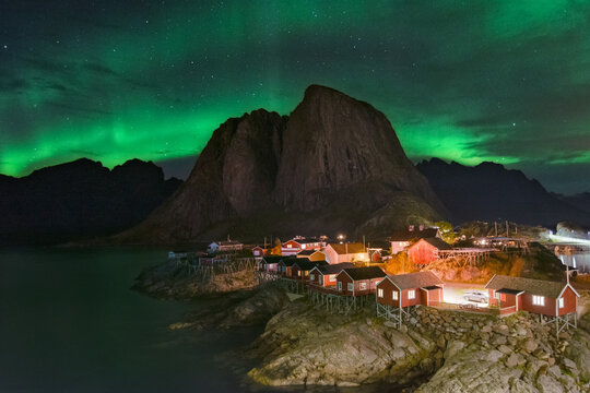 View of the enchanting aurora borealis dances above the quaint red cabins nestled amidst the rugged, rocky landscape, framing the majestic mountains, Lofoten Islands, Nordland, Norway.