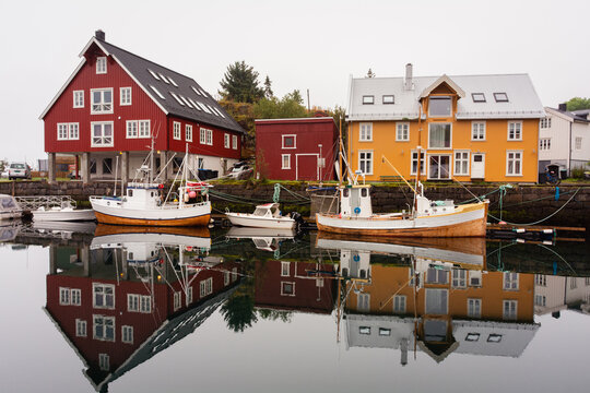 View of vividly colored buildings reflected in the still harbor waters, alongside docked fishing boats, creating a serene yet striking scene, Lofoten Islands, Norway.