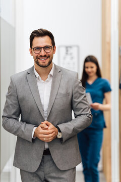 A handsome businessman in a grey suit smiles at the camera while a nurse in blue scrubs reviews charts in the background of a bright, modern healthcare facility.