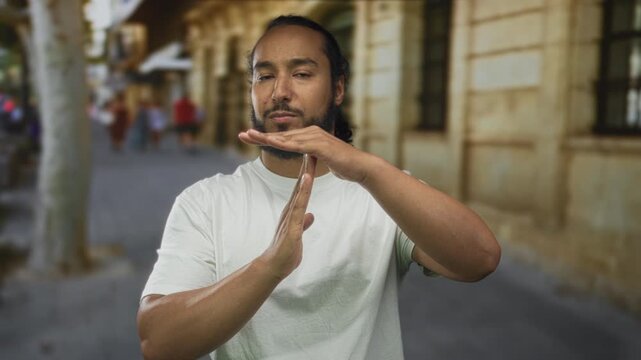Man with beard and white t shirt making timeout gesture with hands on a city street beside stone building facade, pedestrians blurred; assertion.
