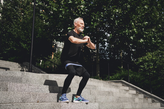 Older bearded man performs a dynamic jump squat on outdoor steps in a park during a morning urban workout