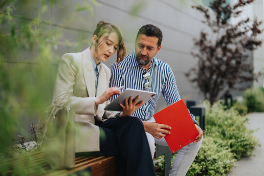Two business colleagues collaborate while reviewing documents on a tablet outdoors. They sit on a bench and discuss work with a red folder during an informal meeting.