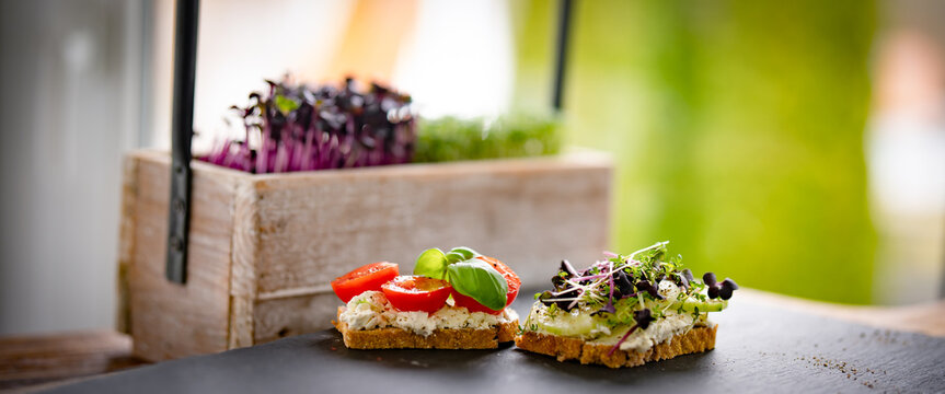 Healthy breakfast bread with cream cheese, cherry tomatoes, basil, cucumber, and fresh cress on a black slate slab. Background for healthy nutrition with space for text. Close-up with short depth of f
