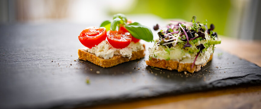 Healthy breakfast bread with cream cheese, cherry tomatoes, basil, cucumber, and fresh cress on a black slate slab. Background for healthy nutrition with space for text.