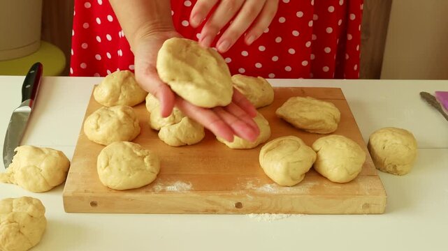 Hands shaping dough into rolls on a wooden board; home baking scene with a red polka-dot apron and fresh buns prep.