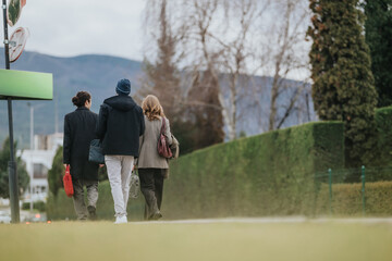 Fototapeta premium Three business associates walk away on a suburban sidewalk carrying bags and coats. A rear view shows colleagues walking together in a landscaped area.