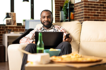 Young black man working remotely with laptop on lap, holding TV remote control while seated on sofa...
