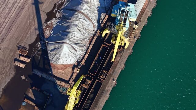 Cranes move material at a busy port. A large pile of cargo sits covered on the dock. Workers coordinate the loading process. Water is visible beside the dock.