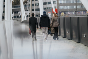 Naklejka premium Three business associates walk away across a modern pedestrian bridge in an urban setting. The colleagues stroll together during a casual city commute, carrying bags and coats.