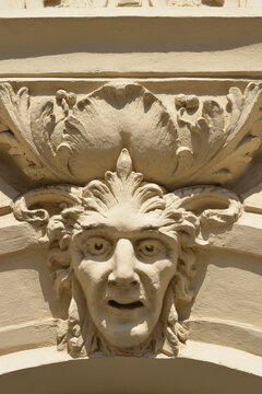 A close up of a decorative stone mascaron depicting a human face with expressive features and acanthus leaf ornaments on a classic building facade.