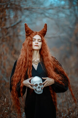 A redhead woman with long fiery hair styled into dramatic horns stands in an autumn forest, holding a skull in her hands. Dressed in a  black dress, she embodies the essence of dark fantasy.  © Valeriia