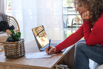 Focused adult woman working on laptop while writing notes on paper at home sofa, managing tasks and...